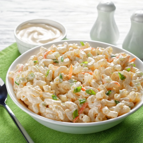 A bowl of homemade macaroni salad with shredded carrot and green onion. In a white bowl on a table sitting on a green table cloth with salt and pepper shakers in the background. 