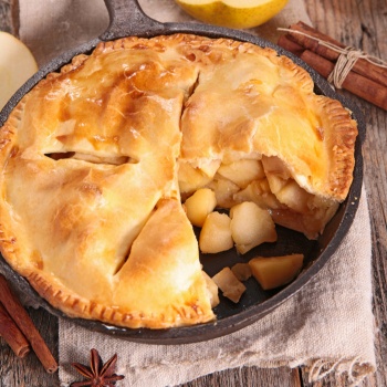 Apple pie baked in a cast iron skillet with a slice cut out. The skillet sits on a kitchen towel on a wooden table with cinnamon sticks and apples in the background. 